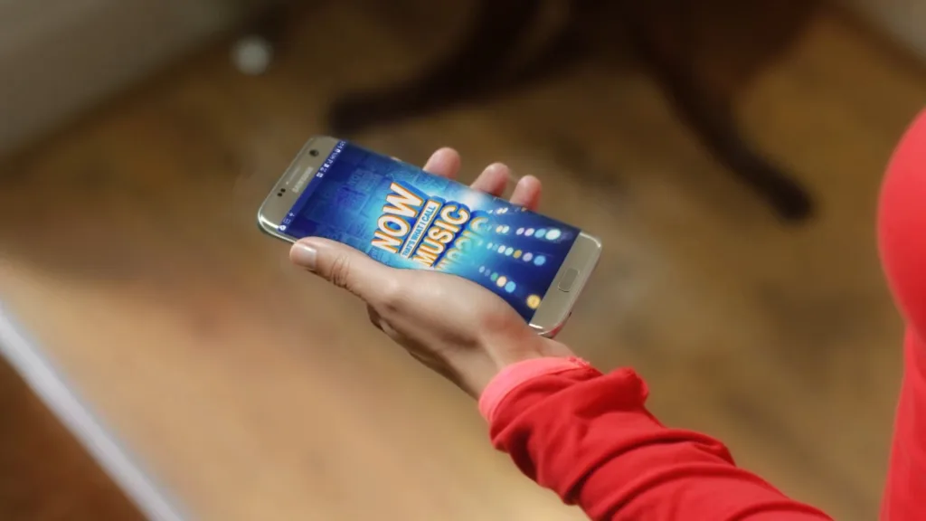 Hand holding a phone showing the NOW Music app