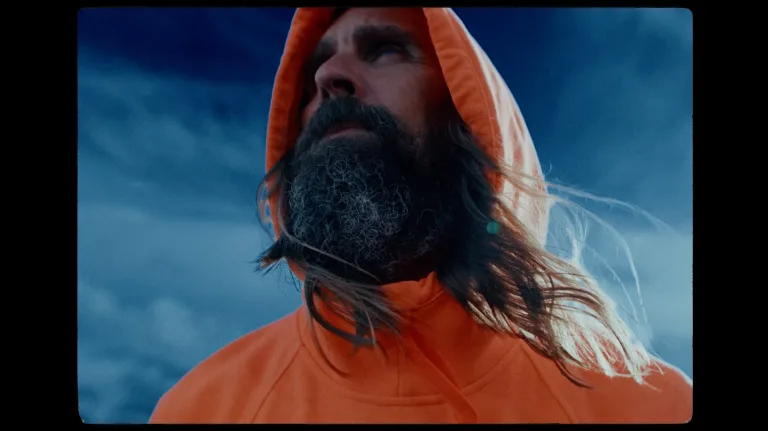 A bearded man in an orange hooded jacket looks up against a stormy blue sky